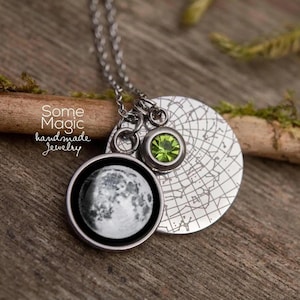 May include: A silver necklace with three charms. One charm is a round pendant with a detailed image of the moon. Another charm is a round pendant with a green gemstone. The third charm is a silver disc with a map design. The text "Some Magic handmade jewelry" is visible.