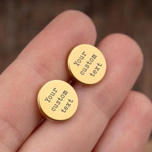 Puede incluir: Dos gemelos redondos dorados con el texto "Your custom text" grabado. Se sostienen entre los dedos, mostrando su superficie pulida y diseño personalizado. El fondo está borroso.