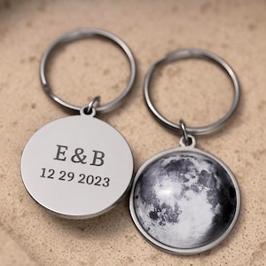 May include: Two silver keychains. One keychain has the initials "E & B" and the date "12 29 2023" engraved on a round pendant. The other keychain features a detailed image of the moon on a round pendant.