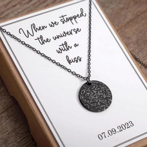 Könnte beinhalten: Eine schwarze Halskette mit einem runden Anhänger, der ein Sternkarten-Design zeigt. Der Anhänger hängt an einer zarten Kette, präsentiert auf einer weißen Karte mit dem Text "When we stopped the universe with a kiss" und dem Datum 07.09.2023.