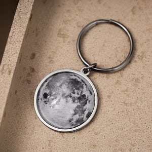 Peut inclure: Un porte-clés rond argenté avec une image détaillée en niveaux de gris de la lune. La surface de la lune est visible avec des cratères et des textures. Le porte-clés est posé sur une surface de couleur neutre.