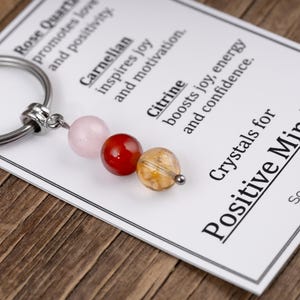 May include: A keychain with three gemstone beads: pink rose quartz, red carnelian, and yellow citrine. The keychain is attached to a card that reads "Crystals for Positive Mindset."