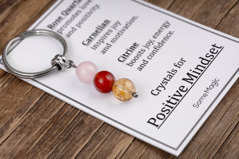 May include: A keychain with three gemstone beads: pink rose quartz, red carnelian, and yellow citrine. The keychain is attached to a card that reads "Crystals for Positive Mindset."