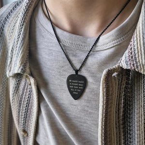 May include: Black guitar pick pendant necklace with the text "We would build a rocket ship and then we'd fly it far away." The pendant hangs on a black chain. The person is wearing a striped jacket and a gray t-shirt.