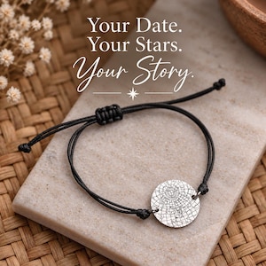 Può includere: Un bracciale in cordino nero con un ciondolo circolare in metallo argentato inciso con una dettagliata mappa stellare. Il testo 'Your Date. Your Stars. Your Story.' è stampato sopra il bracciale in una combinazione di caratteri serif e corsivi.