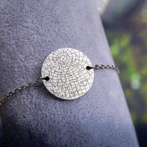 May include: Silver-toned bracelet featuring a circular pendant with a detailed celestial map design. The pendant is connected to a delicate chain. The bracelet is shown against a soft, gray background.