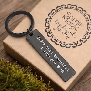 Puede incluir: Un llavero rectangular negro con el texto "Drive safe beautiful. I love you -B" está unido a un anillo negro. El llavero descansa sobre una caja marrón con las palabras "Some Magic handmade Jewelry" en un diseño circular.