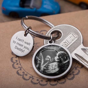 Op de afbeelding: Zilveren sleutelhanger met twee ronde bedels. Eén bedel heeft een echo-afbeelding, de andere zegt "I can't wait to meet you, Daddy!". De sleutelhanger is bevestigd aan een sleutel en een sleutelring.