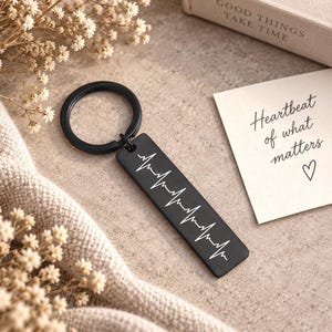 May include: A black rectangular metal keyring featuring a white engraved heartbeat rhythm line. The keyring includes a circular black metal ring. Next to the keyring is a white card with the text Heartbeat of what matters and a heart symbol.