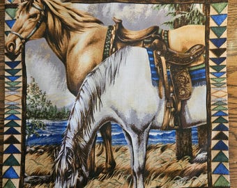 May include: A decorative pillow featuring a colorful geometric border with two horses, one brown and one white, wearing saddles and bridles. The horses are standing in a field with a lake in the background.