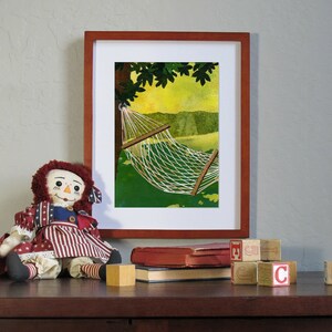 May include: A framed print of a hammock hanging between two trees. The hammock is white and the trees are green. The print is in a wooden frame. A rag doll is sitting in front of the print.