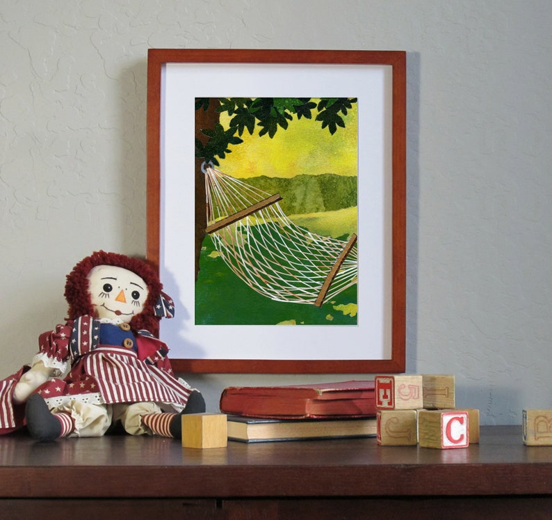 May include: A framed print of a hammock hanging between two trees. The hammock is white and the trees are green. The print is in a wooden frame. A rag doll is sitting in front of the print.