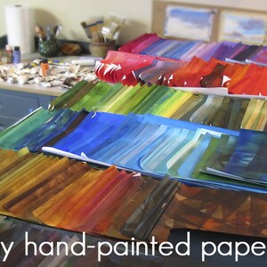 May include: A table covered in stacks of hand-painted papers in various colours, including red, orange, yellow, green, blue, and purple. The text "my hand-painted papers" is visible at the bottom of the image.