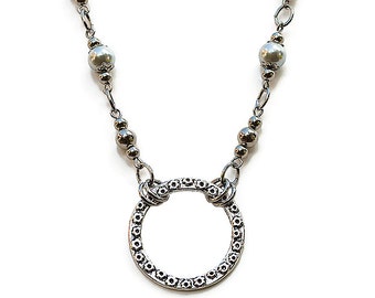 May include: A silver necklace with a large, circular pendant and two white pearl accents. The pendant has a textured, ornate design.