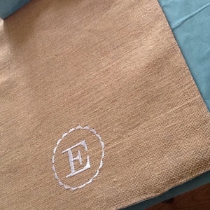 May include: A beige burlap table runner with a white embroidered circle containing the letter "E".