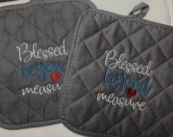 May include: Two gray quilted pot holders with the embroidered text "Blessed beyond measure". The text is in white and blue, with a small red heart. Each pot holder has a loop for hanging.