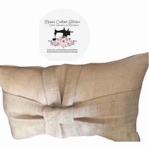 May include: A beige burlap pillow with a large bow in the center. The pillow is made of a natural fabric with a woven texture.