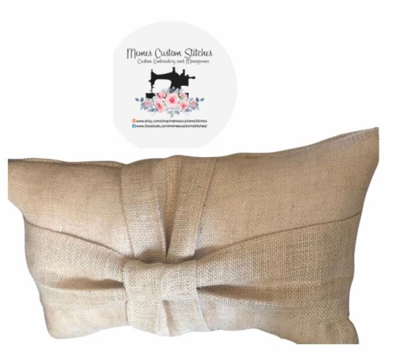 May include: A beige burlap pillow with a large bow in the center. The pillow is made of a natural fabric with a woven texture.