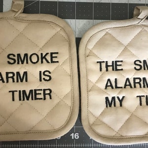May include: Two beige quilted pot holders with black embroidered text that reads "The smoke alarm is my timer".