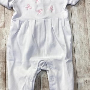 May include: White baby romper with pink bow embroidery. The romper features a Peter Pan collar with the name "Charlie" embroidered in pink. The garment has short sleeves and snaps for easy closure. A tag is attached to the collar.