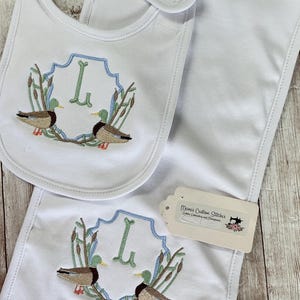 May include: White baby bib and burp cloth set. Each item features an embroidered design of two ducks, reeds, and the letter "L" in a blue and green frame. The bib has a neck strap. A tag with text is attached.
