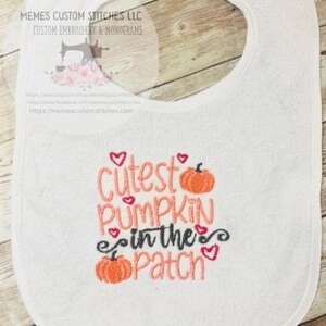 May include: White baby bib with the embroidered phrase "Cutest Pumpkin in the Patch" in orange and black thread, with small orange pumpkin and heart accents. The bib has a rounded shape.