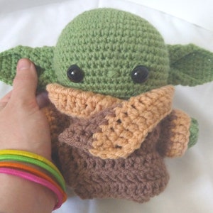 Pu&ograve; includere: Un giocattolo di peluche verde e marrone all'uncinetto con grandi occhi. Il giocattolo indossa una sciarpa all'uncinetto marrone e arancione.
