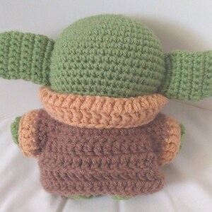 Pu&ograve; includere: Un giocattolo di peluche all'uncinetto che ricorda un personaggio del franchise di Star Wars. Il giocattolo &egrave; verde con grandi orecchie e un vestito marrone e arancione.