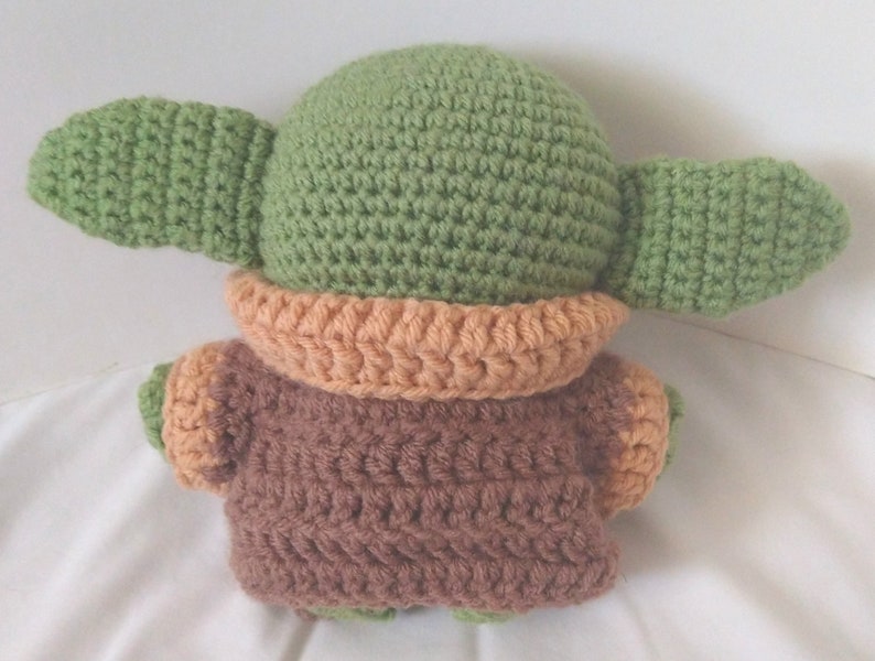 Pu&ograve; includere: Un giocattolo di peluche all'uncinetto che ricorda un personaggio del franchise di Star Wars. Il giocattolo &egrave; verde con grandi orecchie e un vestito marrone e arancione.