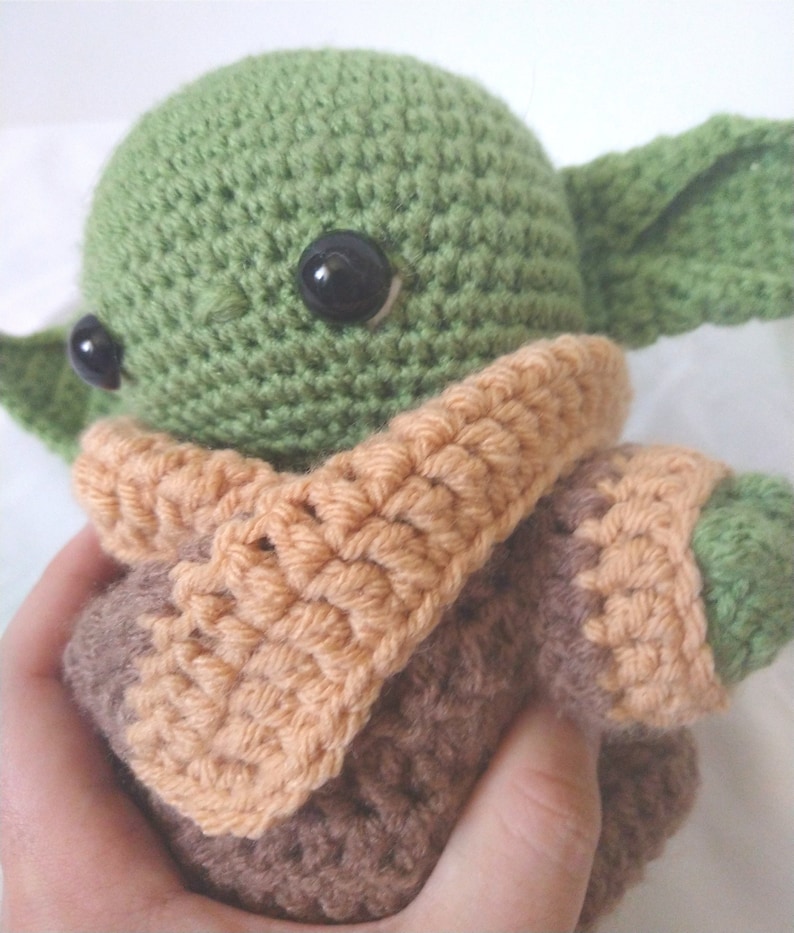 Pu&ograve; includere: Un giocattolo di peluche verde e marrone all'uncinetto con grandi occhi neri. Il giocattolo indossa una sciarpa all'uncinetto marrone e arancione.