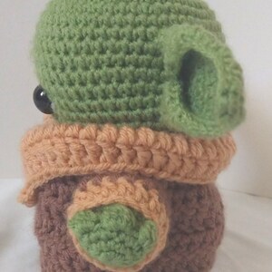 Pu&ograve; includere: Un giocattolo di peluche all'uncinetto a forma di personaggio verde con grandi orecchie, vestiti marroni e arancioni e occhi neri.