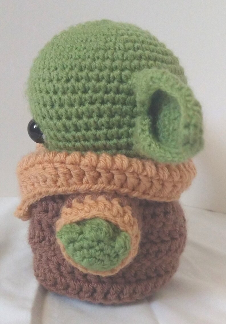 Pu&ograve; includere: Un giocattolo di peluche all'uncinetto a forma di personaggio verde con grandi orecchie, vestiti marroni e arancioni e occhi neri.