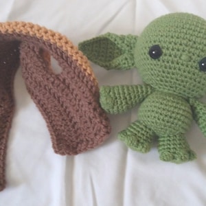 Pu&ograve; includere: Un giocattolo di peluche verde all'uncinetto che ricorda un personaggio di Star Wars, con grandi occhi neri. Il giocattolo &egrave; seduto accanto a una sciarpa all'uncinetto marrone e arancione.