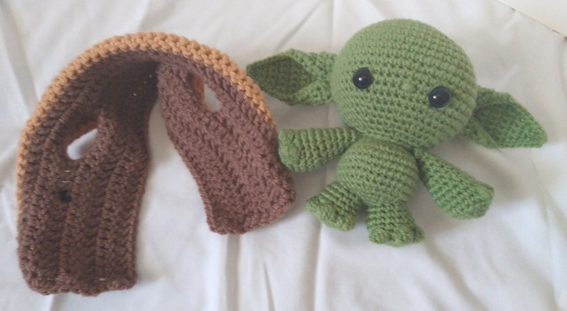 Pu&ograve; includere: Un giocattolo di peluche verde all'uncinetto che ricorda un personaggio di Star Wars, con grandi occhi neri. Il giocattolo &egrave; seduto accanto a una sciarpa all'uncinetto marrone e arancione.