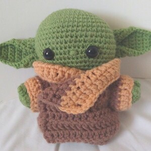Pu&ograve; includere: Un giocattolo di peluche verde all'uncinetto con occhi grandi e vestiti marroni e arancioni. Il giocattolo &egrave; un personaggio popolare del franchise di Star Wars.