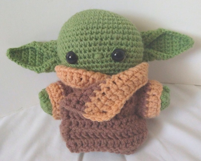 Pu&ograve; includere: Un giocattolo di peluche verde all'uncinetto con occhi grandi e vestiti marroni e arancioni. Il giocattolo &egrave; un personaggio popolare del franchise di Star Wars.