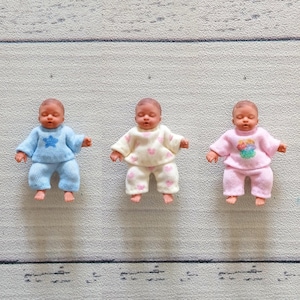 May include: Three miniature baby dolls wearing knitted outfits. The first doll wears a light blue jumper with a blue star and matching trousers. The second doll wears a cream jumper with pink hearts and matching trousers. The third doll wears a pink jumper with a colourful graphic and matching trousers.