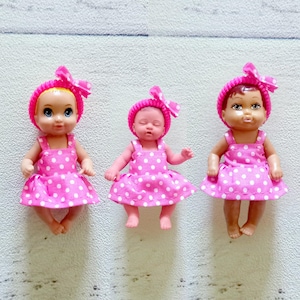 May include: Three miniature baby dolls. Each doll wears a pink dress with white polka dots and a matching pink knit cap with a bow. The dolls have different skin tones and facial expressions, with one doll appearing to be sleeping.