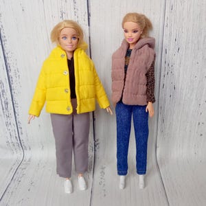 Könnte beinhalten: Zwei Puppen in modischen Outfits. Eine Puppe trägt eine gelbe Steppjacke, graue Hose und weiße Turnschuhe. Die andere Puppe hat eine rosa Weste, blaue Jeans und weiße Schuhe. Beide Puppen haben blondes Haar und stehen vor hellem Holz.