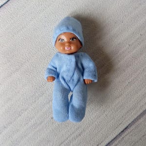May include: A small doll wearing a light blue knit hat and matching onesie. The doll has brown skin, a pink mouth, and detailed facial features. The doll is standing upright, showcasing the full outfit.