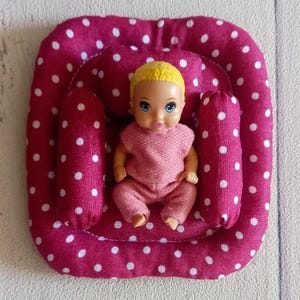 May include: A doll in a pink outfit sits in a pink and white polka dot baby seat. The doll has blonde hair and blue eyes. The seat has two side bolsters and a rounded base.