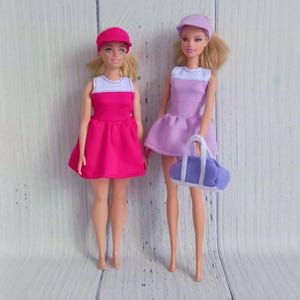 May include: Two dolls wearing matching outfits. One doll is in a pink dress and cap, the other in a purple dress and cap, holding a purple and white bag. Both dresses have a white top.