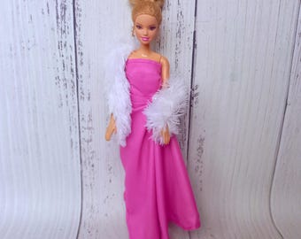 Può includere: Una bambola con un abito rosa acceso senza spalline e una stola di pelliccia sintetica bianca. La bambola ha i capelli biondi raccolti in uno chignon e piccoli orecchini. L'abito è lungo e ha un design drappeggiato.