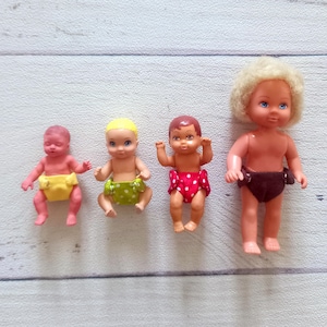 Könnte beinhalten: Vier Vintage-Babypuppen in verschiedenen Größen. Die Puppen haben unterschiedliche Hauttöne und tragen Windeln in Gelb, Grün mit weißen Punkten, Rot mit weißen Punkten und Braun. Die Puppen haben blonde, braune und hellrosa Haare.
