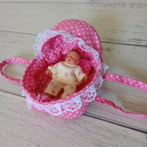 Puede incluir: Una muñeca en miniatura de tez bronceada que viste una camiseta de manga larga y pantalones de color crema con un patrón de corazones rosas, descansando dentro de un pequeño moisés de tela rosa con lunares blancos y encaje blanco.