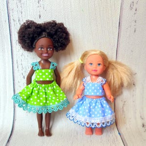May include: Two small dolls are shown, one with dark skin and the other with fair skin. Both dolls wear matching outfits: a crop top and skirt with white polka dots. The dark-skinned doll's outfit is green, and the fair-skinned doll's is blue.