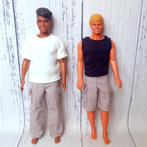 May include: Two fashion dolls are displayed. One doll wears a white t-shirt and light gray pants. The other doll is dressed in a black tank top and light gray cargo shorts. Both dolls have a tan skin tone and are standing.