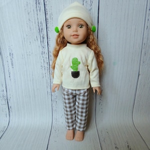 May include: A doll with long wavy reddish-brown hair wearing a cream-colored beanie with small green pom-poms, a cream long-sleeve shirt featuring a green cactus graphic in a dark brown pocket, and brown and white checkered pants.