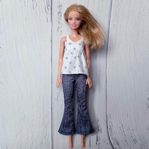 May include: A doll wearing a white tank top with blue floral designs and dark blue denim bell-bottom pants. The doll has blonde hair and is posed against a white wooden background. The tank top has a scoop neck.