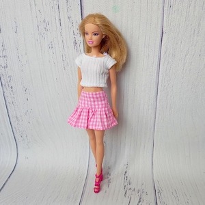 May include: A doll wearing a white, short-sleeved crop top and a pink and white checkered skirt. The doll is also wearing pink high heels. The doll has blonde hair and is standing in front of a white wooden background.